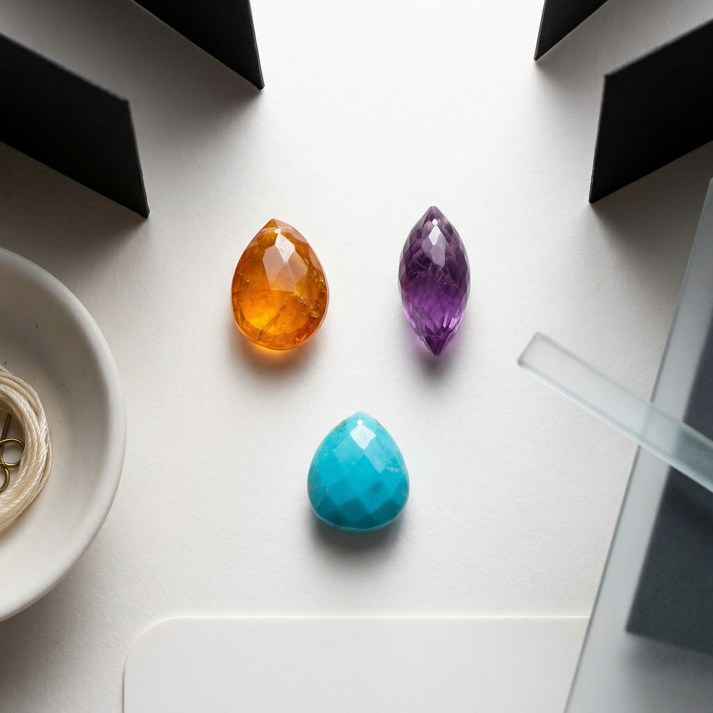 Three briolette gems on warm white paper with porcelain bowl and frosted glass leading line.