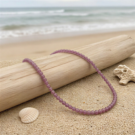 Single strand of tiny faceted rhodolite garnet beads resting on a neutral surface, rich wine-red facets catching soft, natural studio light.