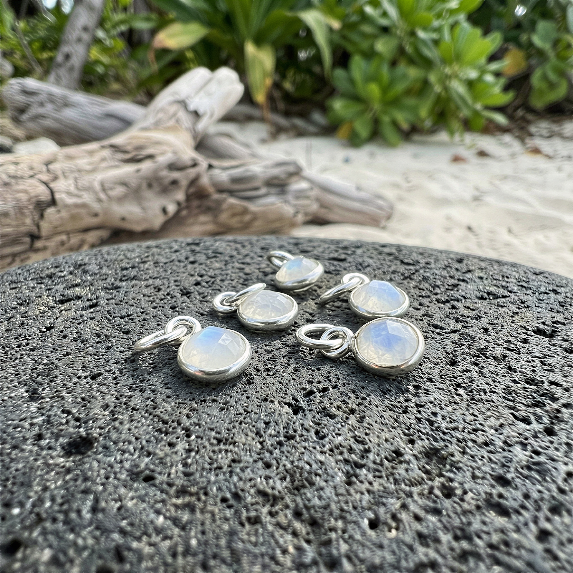 Rainbow moonstone pendant in sterling silver bezel resting on sunlit volcanic stone with tropical foliage.