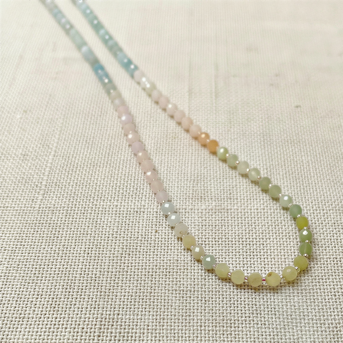 Soft mixed beryl beads in gentle pastel hues drape as a continuous strand on textured linen, lit with airy natural light.