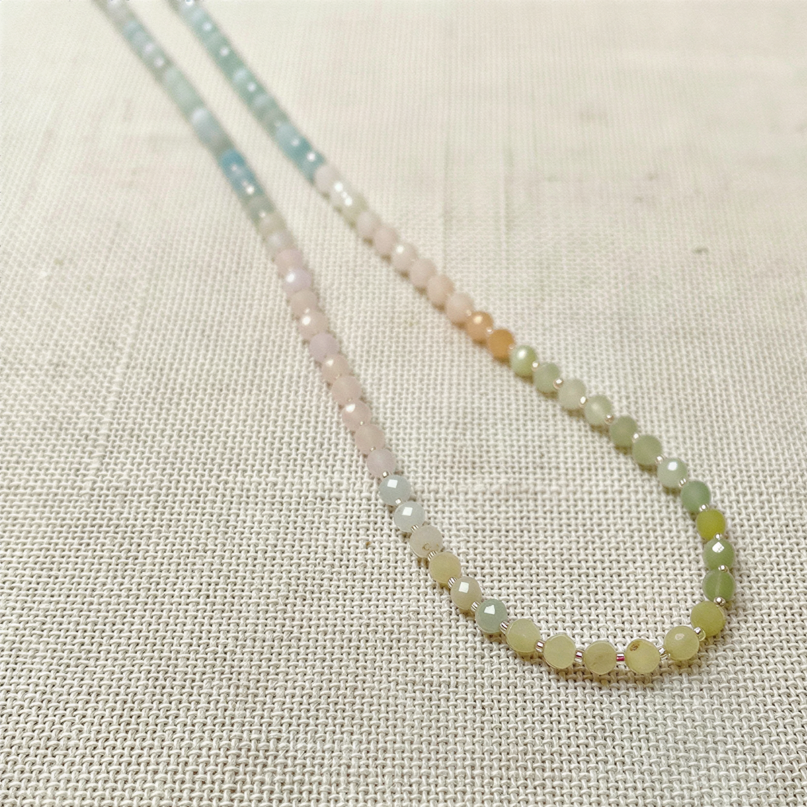 Soft mixed beryl beads in gentle pastel hues drape as a continuous strand on textured linen, lit with airy natural light.
