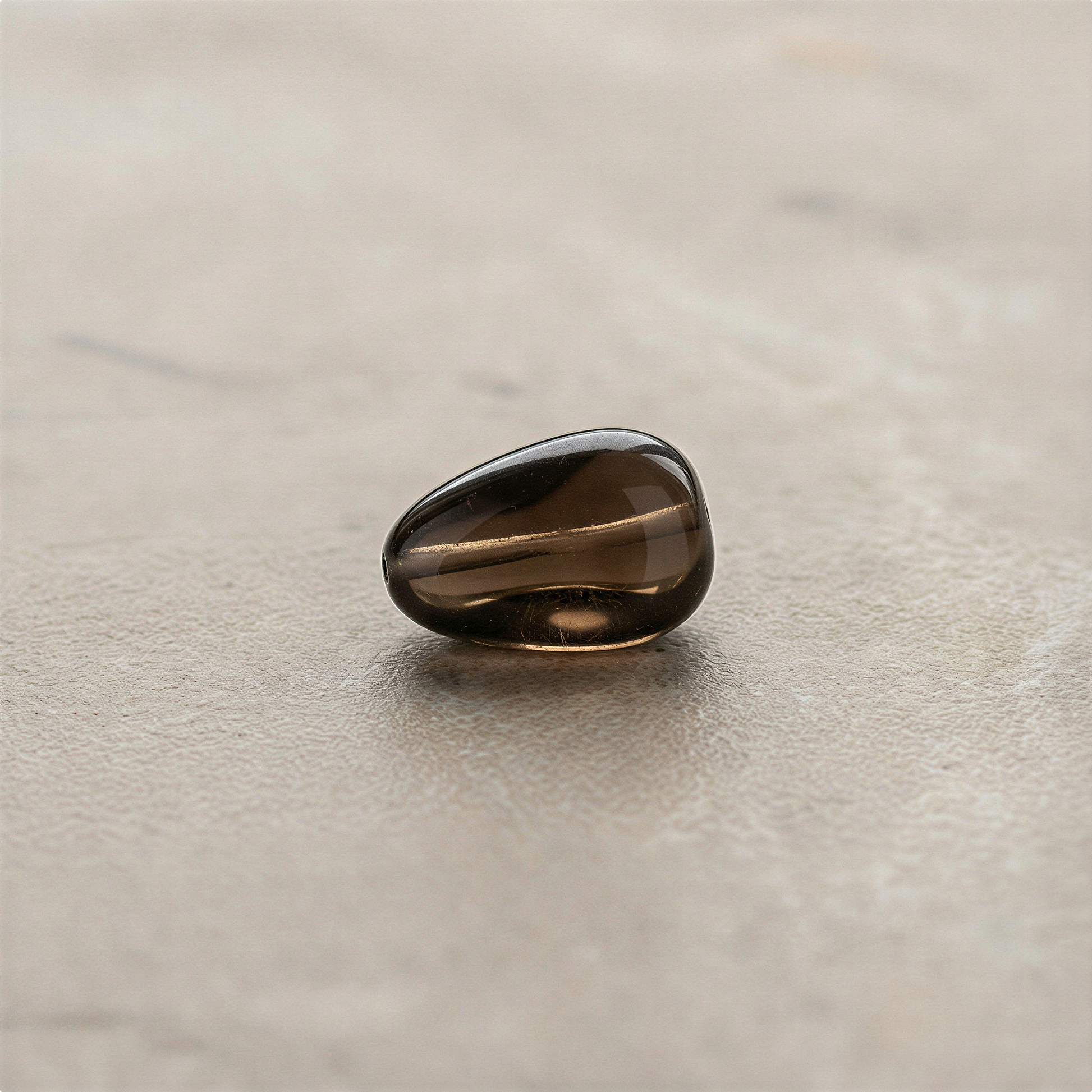 Smoky quartz tumbled nugget bead displayed on neutral surface with soft studio lighting enhancing natural depth