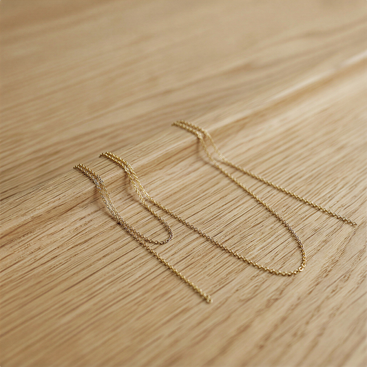 Two delicate gold-filled micro cable chains draped softly over a warm oak-toned surface, catching diffused morning light with subtle organic texture beneath.
