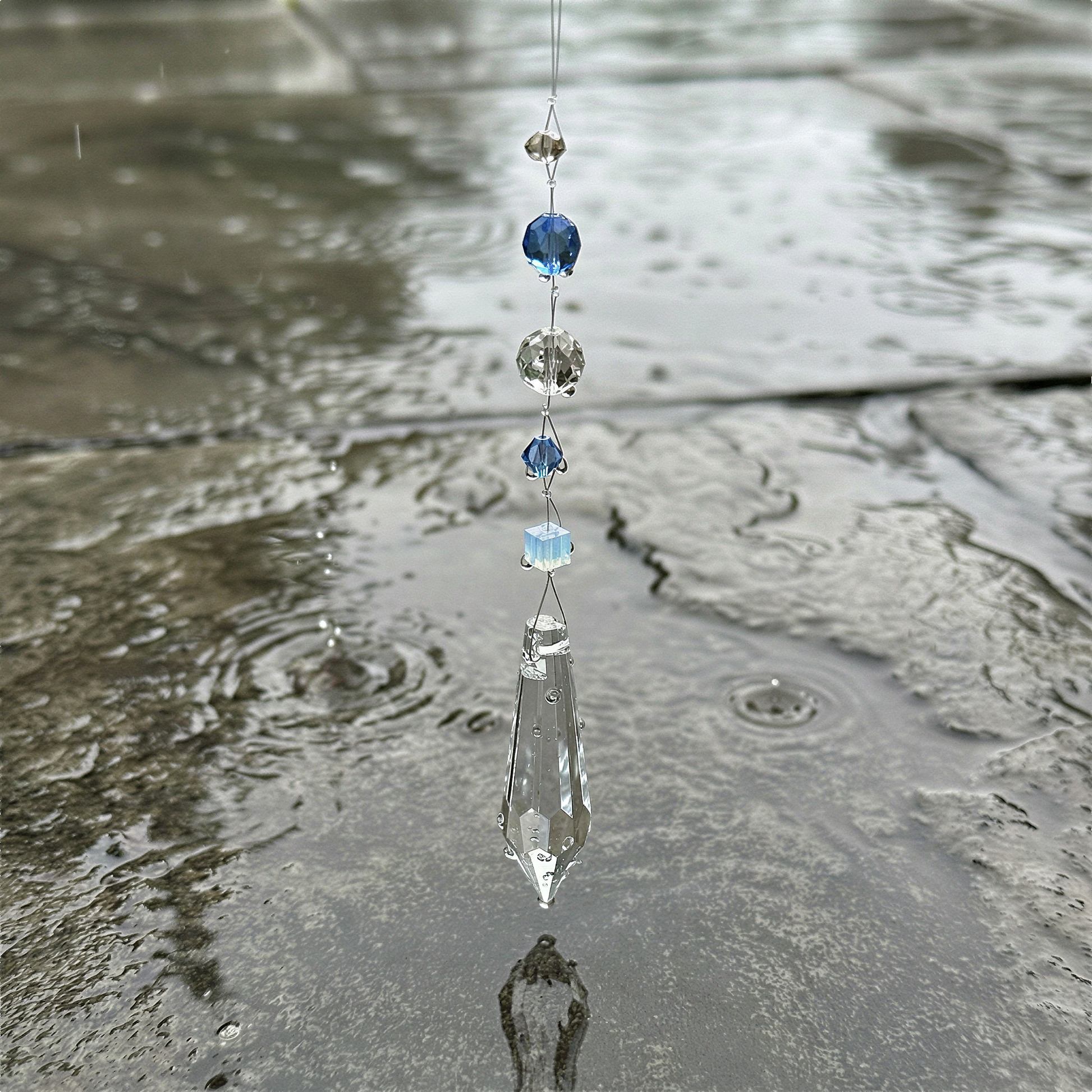 Crystal lightcatcher ornament resting near window, catching soft daylight with droplets and tranquil summer rain ambiance