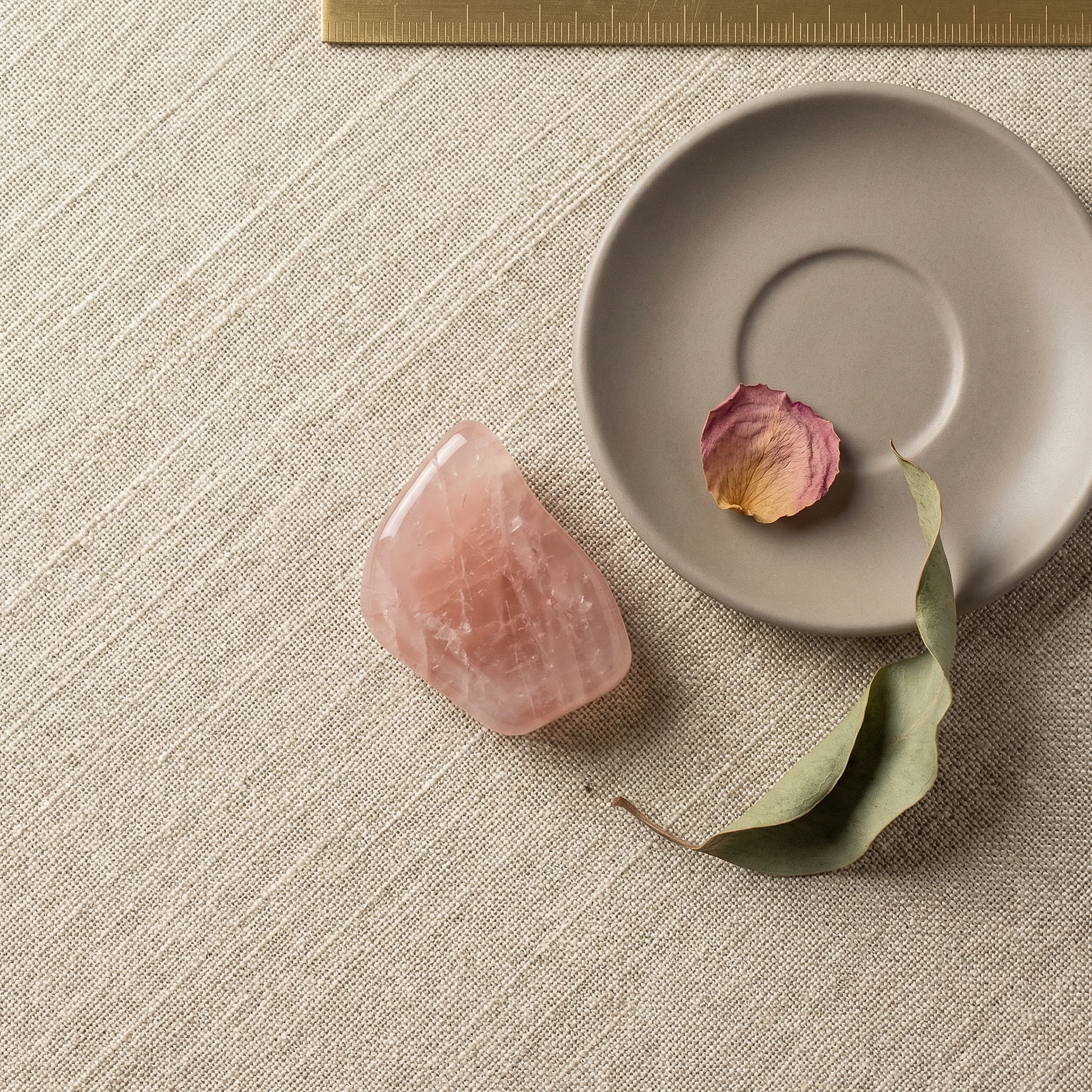 Overhead rose quartz on oatmeal linen with grey saucer, dried petal, and brass rule