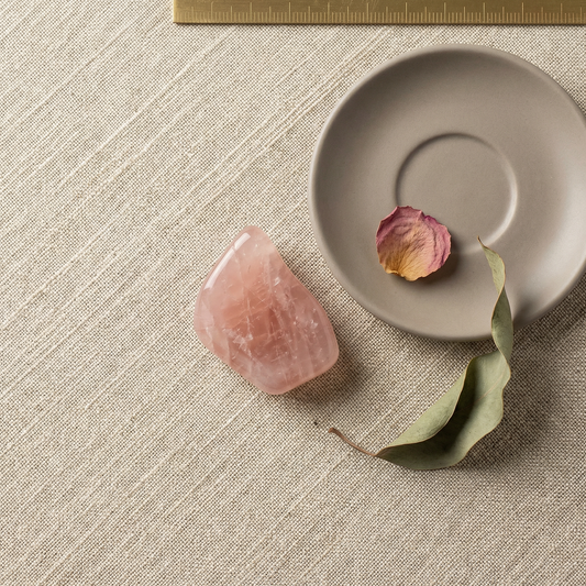 Overhead rose quartz on oatmeal linen with grey saucer, dried petal, and brass rule