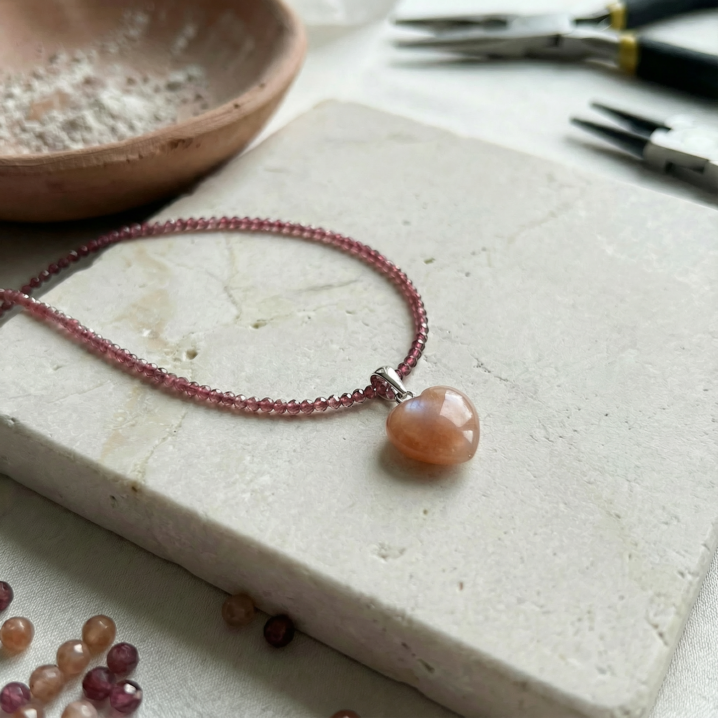 Luminous peach moonstone heart pendant with soft translucence, suspended on a faceted gemstone necklace, resting on a textured stone surface in gentle studio light.