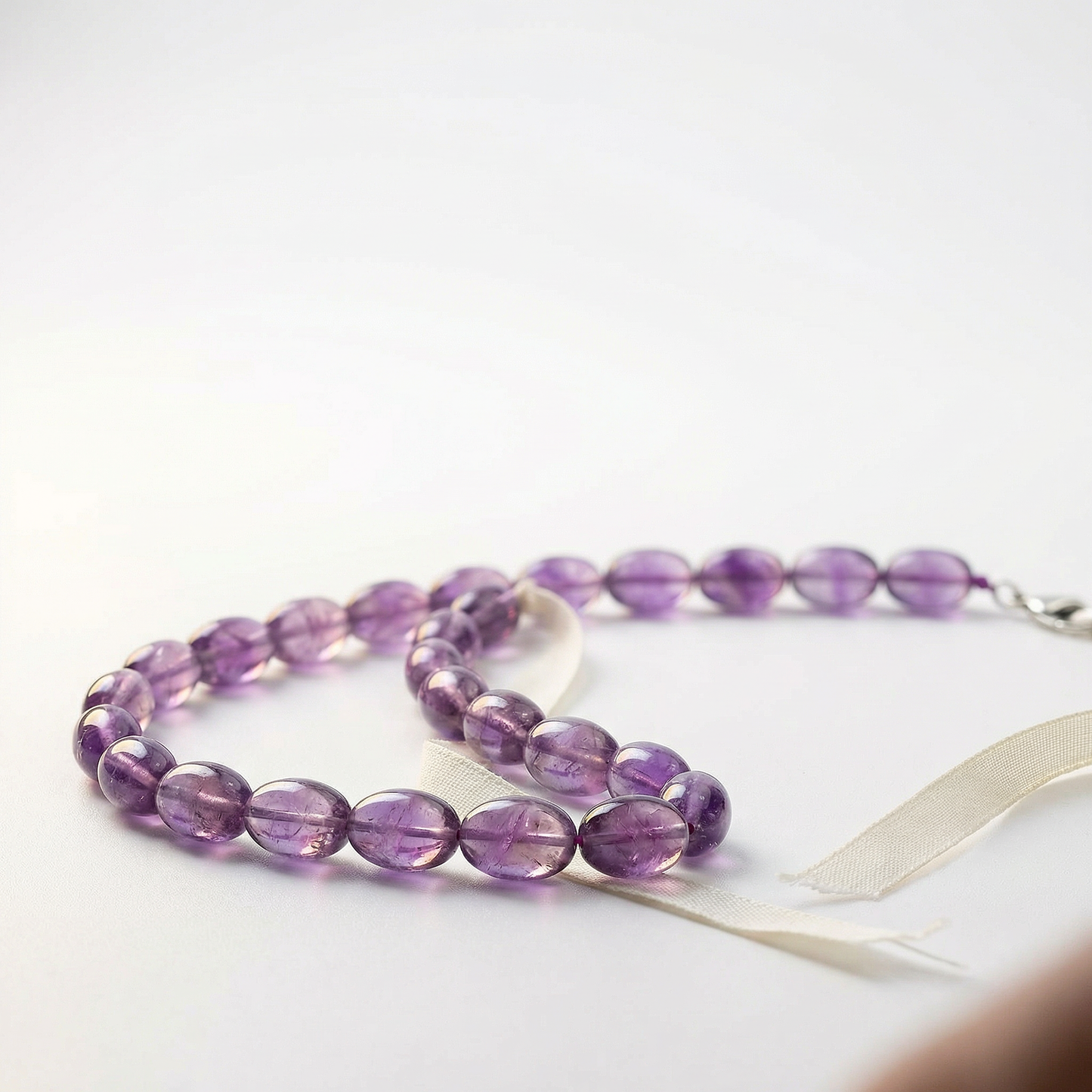 Amethyst bead strand on white matte surface with linen ribbon and warm rim light.