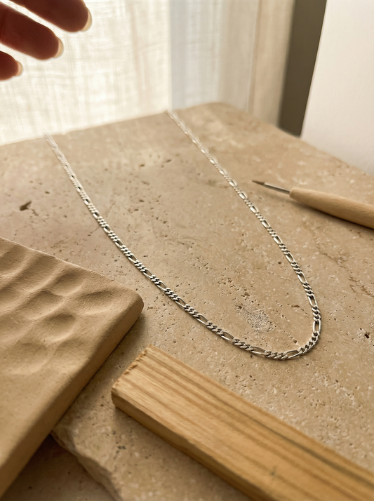 18" small Figaro silver chain draped on warm limestone with clay and olive-wood accents.