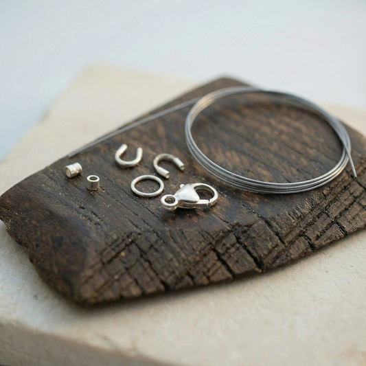 Silver-tone jewelry findings arranged on a dark wooden block, paired with a thin steel-gray beading cable softly coiled under gentle studio light